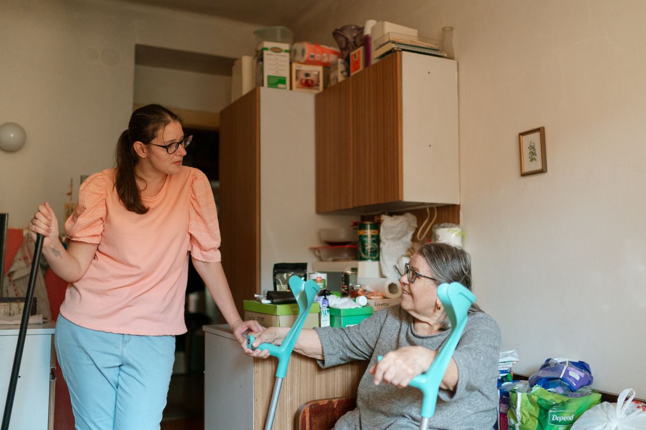 Services A caregiver engaging with a senior using crutches in a cozy Prague home environment.