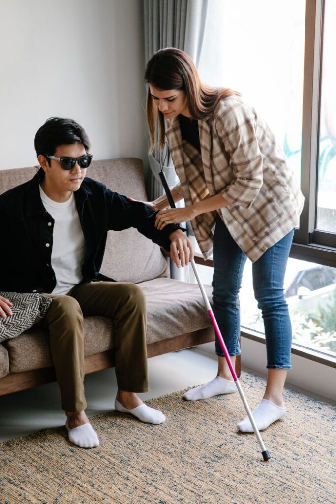 Home A woman provides guidance to a visually impaired man using a walking stick indoors on a cozy sofa.