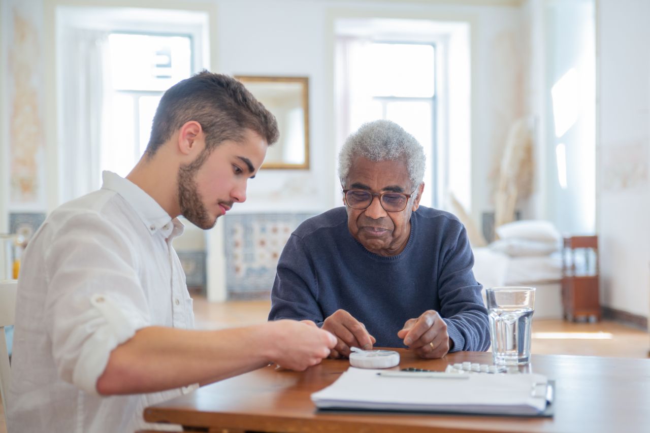Services Young man assisting elderly man with medication in a bright living room setting.