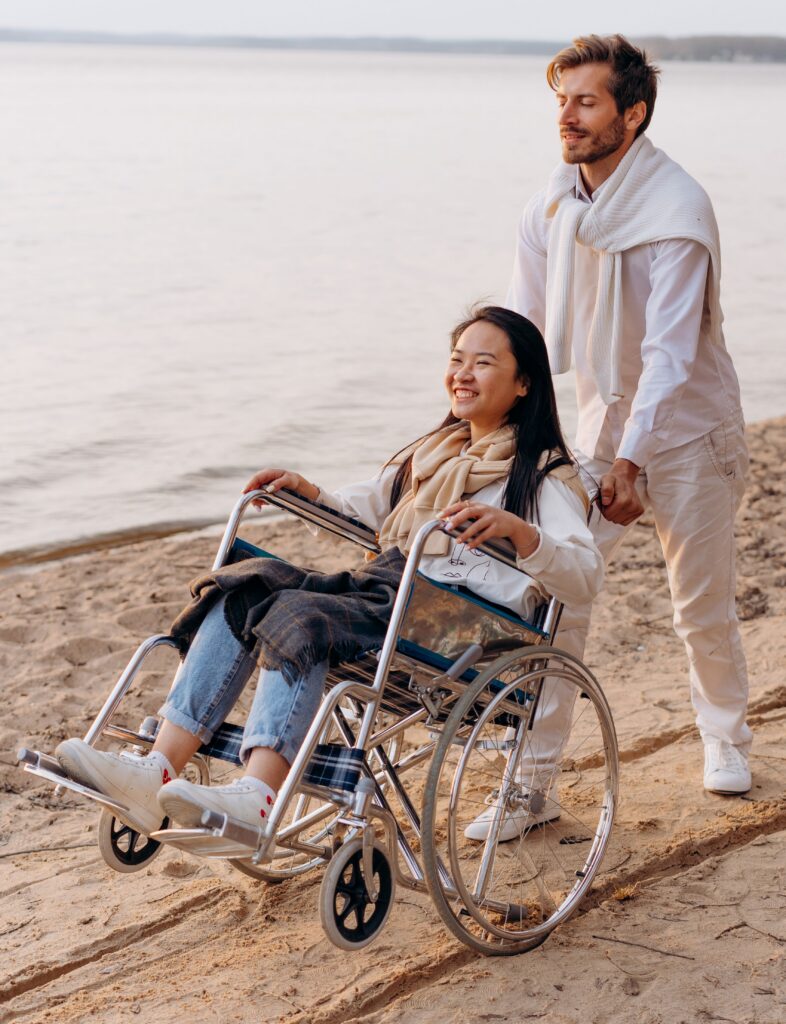 Home A happy couple at the beach, with a man pushing a woman in a wheelchair along the shore.